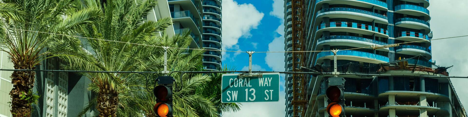 A street sign for Coral Way SW 13 St, traffic lights, palm trees, and modern high-rise buildings.