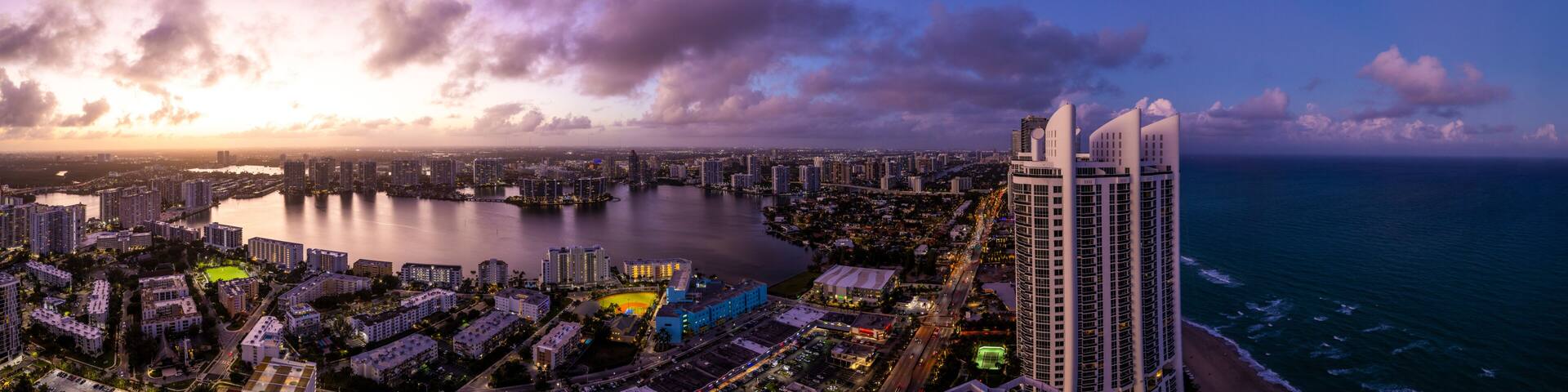 A coastal cityscape at dusk with a prominent modern skyscraper, a marina, and a densely built urban area.