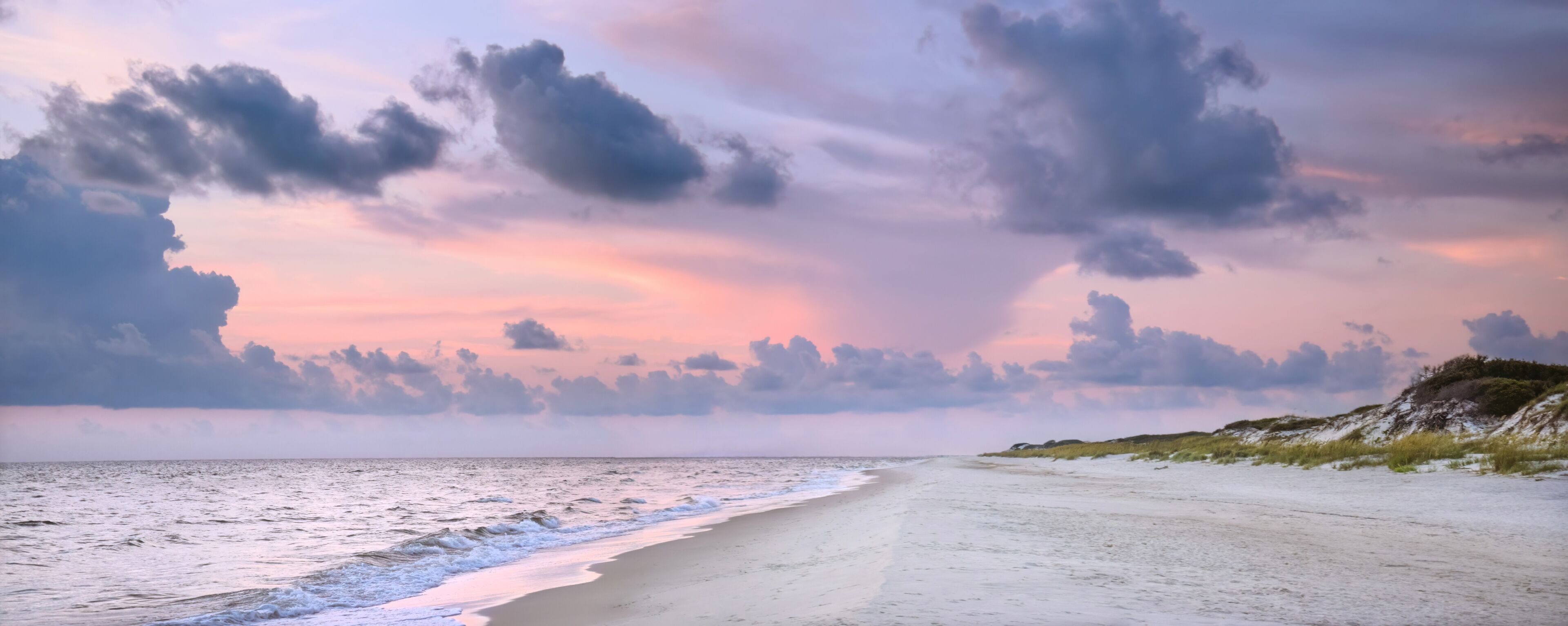 A beach with a sandy shore and waves, under a cloudy sky with pink and purple hues.