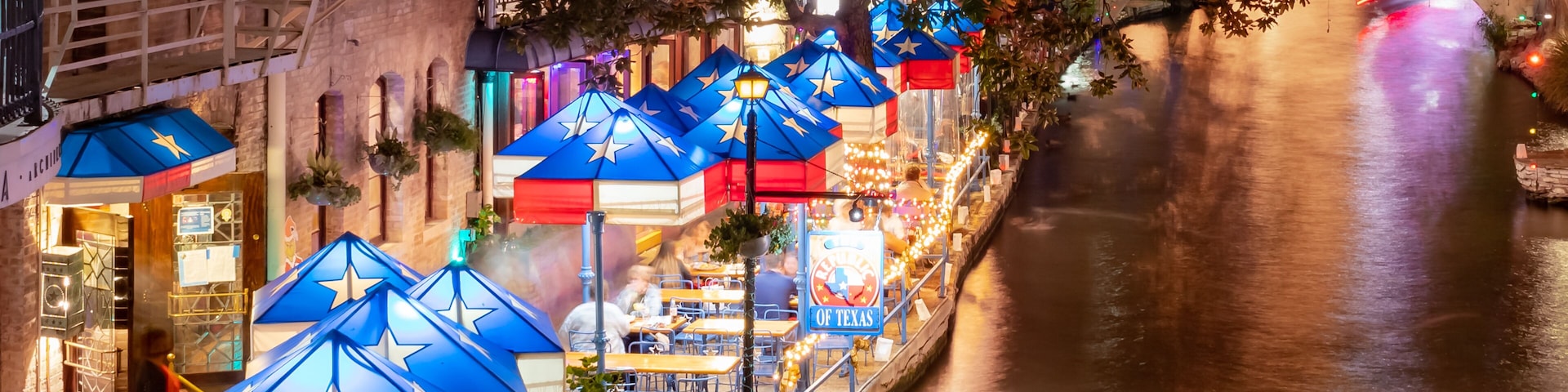 A riverside walkway with illuminated blue and red umbrellas, a sign reading 'Republic of Texas,' and a brick building with a fire escape.