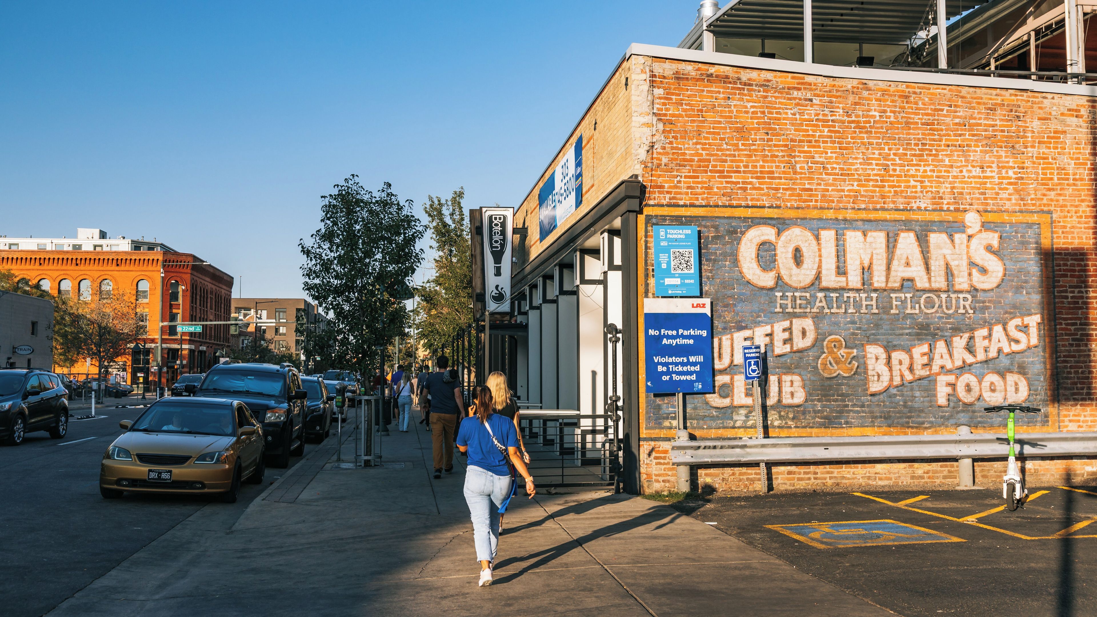 Une rue bordée de voitures garées, un bâtiment en briques avec une grande enseigne indiquant « Colman's Health Flour Club », et un piéton qui passe.