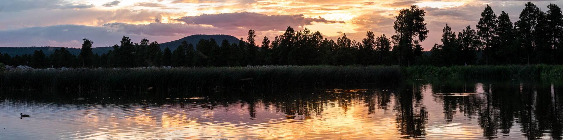A sunset over a calm lake with reflections of the sky, surrounded by a forest of tall trees.