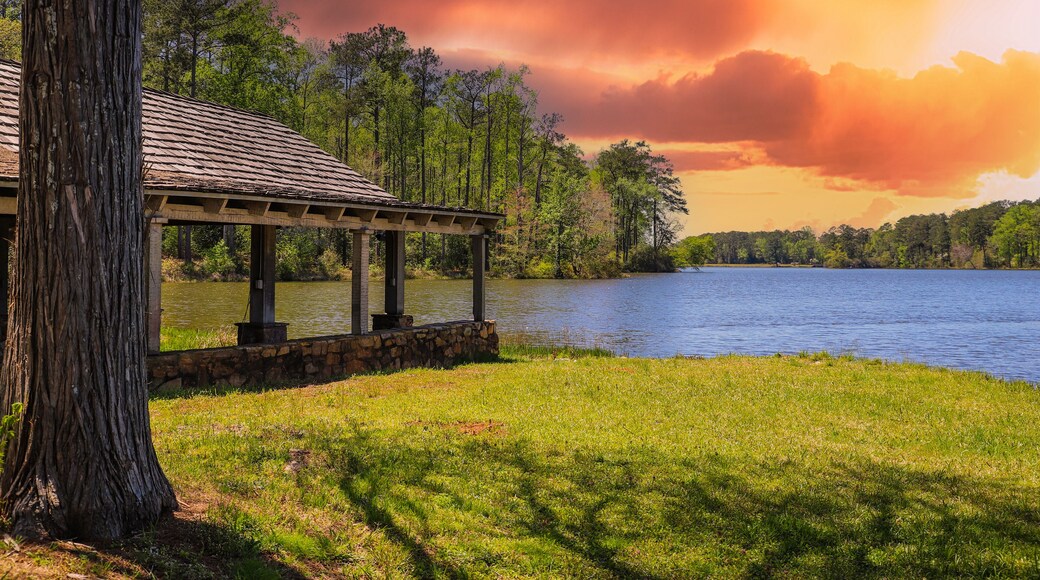 A brown wooden boat house on a gorgeous vast blue lakes surrounded by lush green trees, grass and plants with powerful red clouds at sunset at Callaway Gardens in Pine Mountain Georgia USA