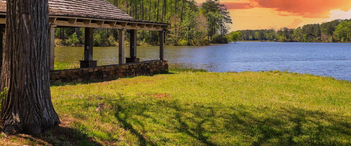 A brown wooden boat house on a gorgeous vast blue lakes surrounded by lush green trees, grass and plants with powerful red clouds at sunset at Callaway Gardens in Pine Mountain Georgia USA