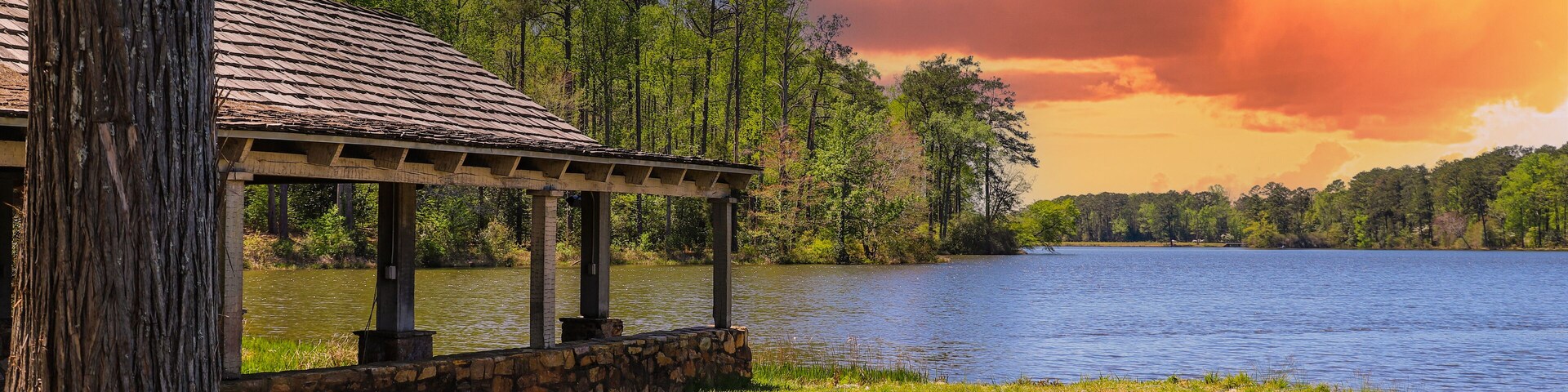 A brown wooden boat house on a gorgeous vast blue lakes surrounded by lush green trees, grass and plants with powerful red clouds at sunset at Callaway Gardens in Pine Mountain Georgia USA