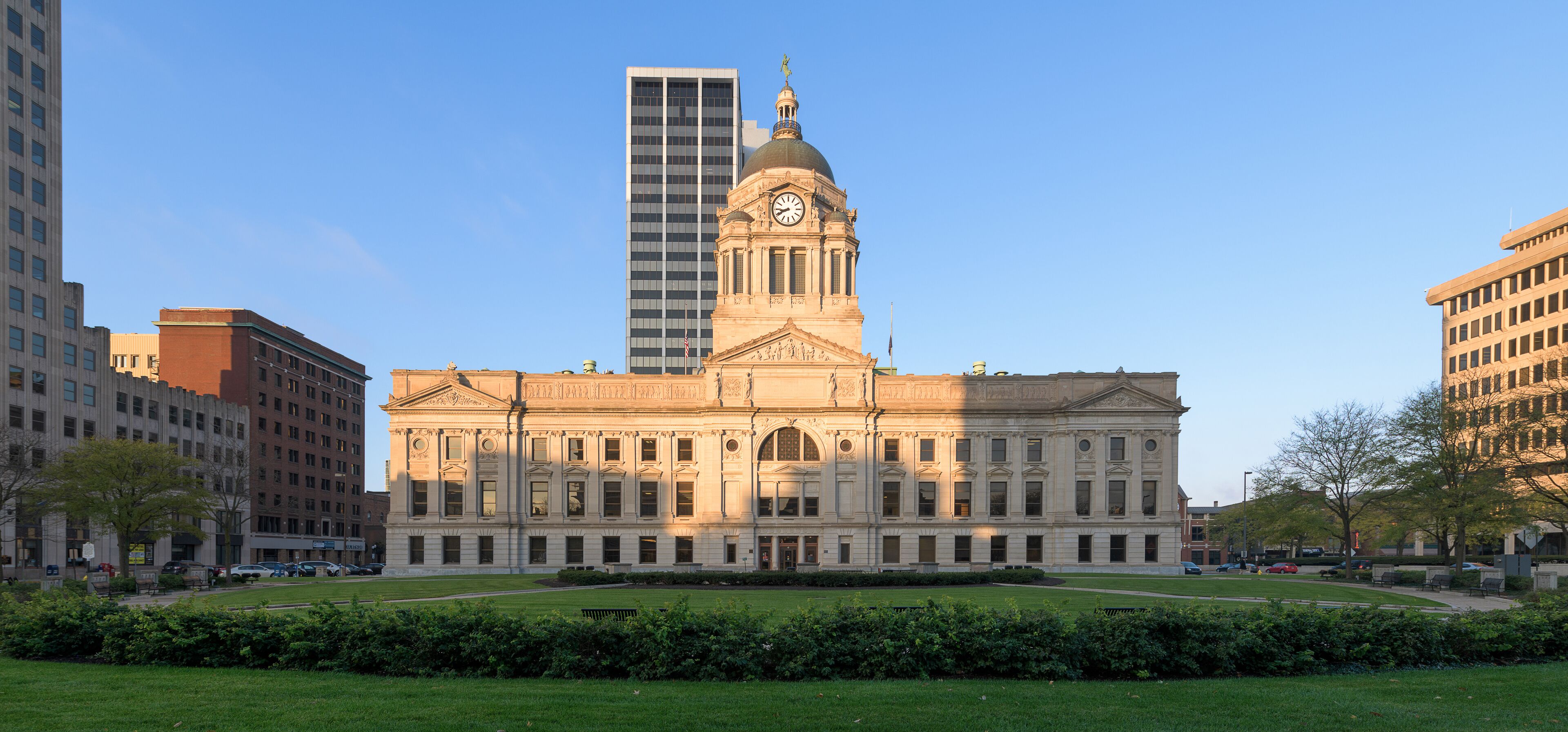 A historic building with a clock tower, surrounded by modern skyscrapers and a well-maintained lawn.