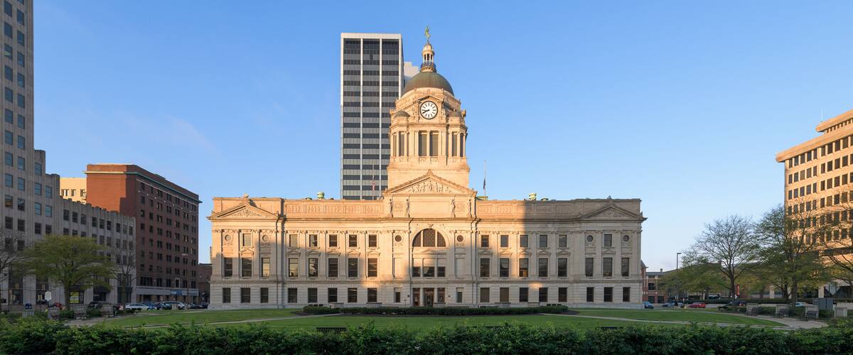 A historic building with a clock tower, surrounded by modern skyscrapers and a well-maintained lawn.