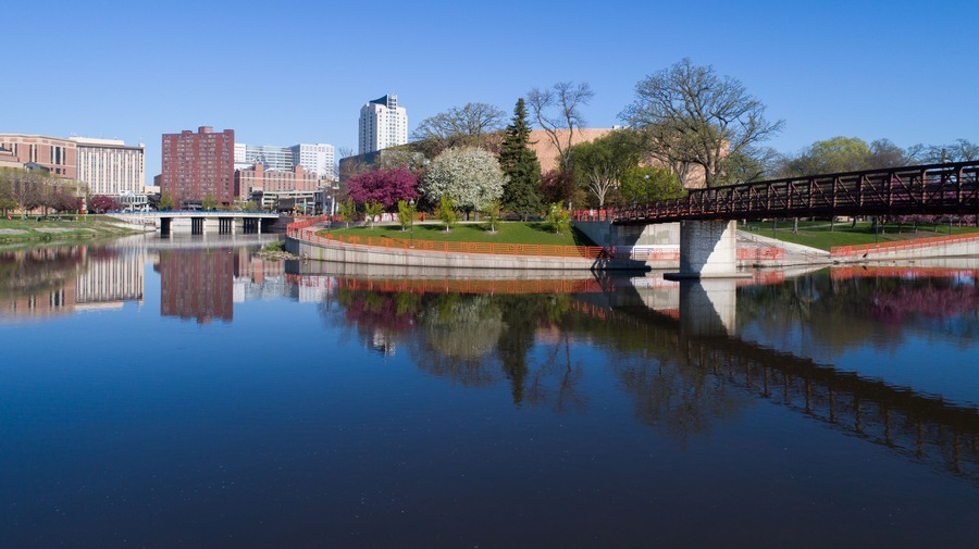 A calm river reflecting a bridge and buildings, surrounded by trees and a clear sky.