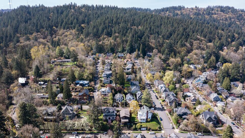 Aerial drone view of Northwest Portland, Oregon, showing Forest Park, Nob Hill, and Willamette Heights with greenery, homes, and industrial areas along the Willamette River