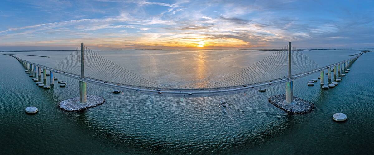 A large suspension bridge over calm water with a sunset in the background.