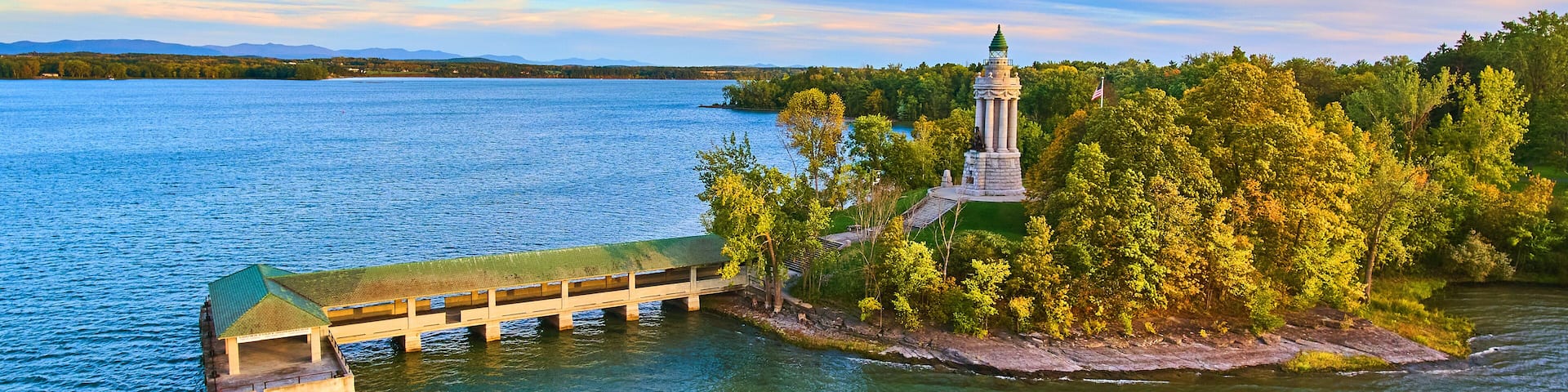 A wooden pier extending into a lake, surrounded by lush greenery and a prominent lighthouse on a small island.