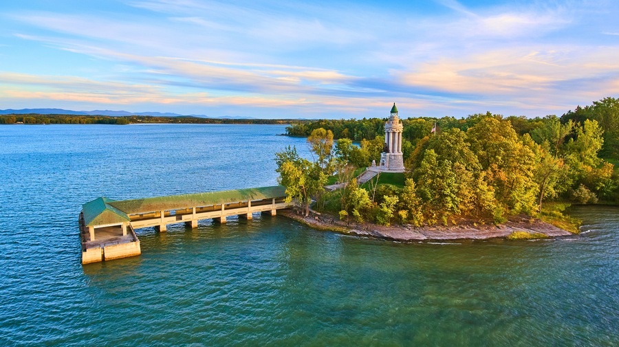 A wooden pier extending into a lake, surrounded by lush greenery and a prominent lighthouse on a small island.
