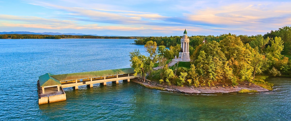 A wooden pier extending into a lake, surrounded by lush greenery and a prominent lighthouse on a small island.