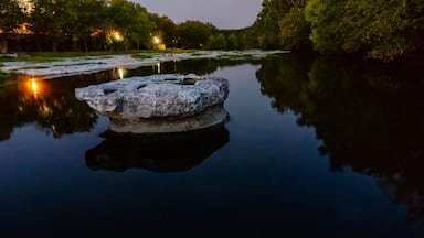 Um rio calmo com uma rocha grande ao centro, rodeado por árvores e iluminado por luzes distantes.