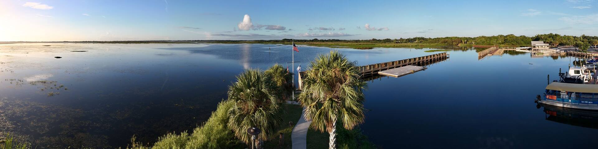 A calm river with a wooden dock extending into the water, surrounded by lush greenery and a clear sky.