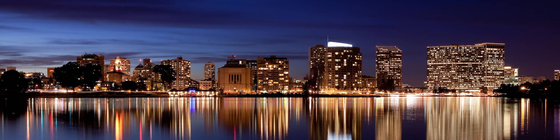 Un horizonte urbano nocturno con edificios iluminados reflejados en aguas tranquilas.