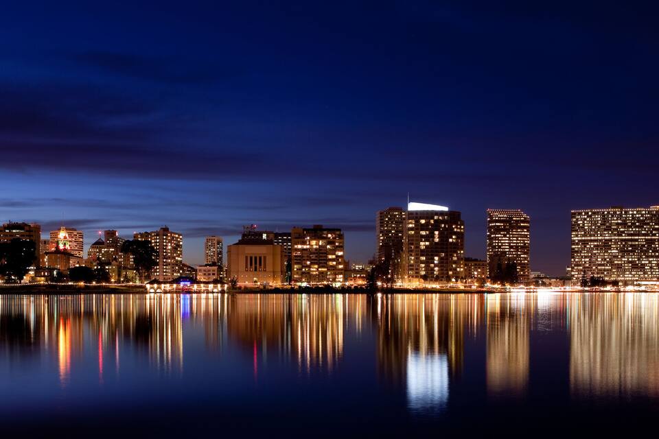 Un horizonte urbano nocturno con edificios iluminados reflejados en aguas tranquilas.