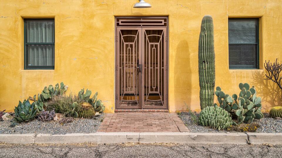 A yellow building with a wooden door, cacti, and a tall cactus in the garden.