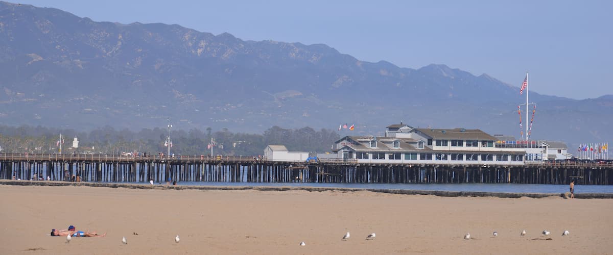 A beach with a pier extending into the water, mountains in the background, and several flags on the pier.