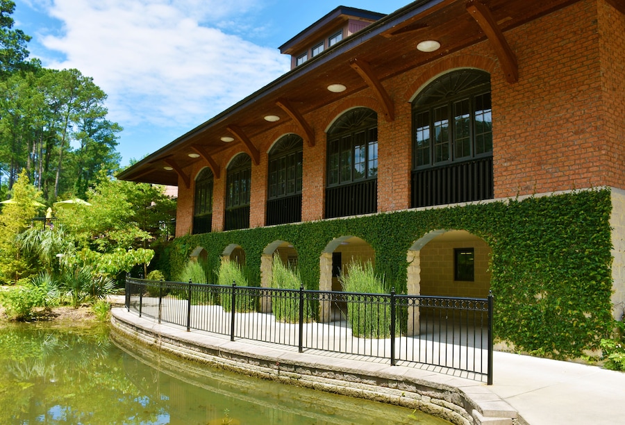 Un bâtiment historique doté d’arcs en plein cintre et d’une façade en briques, situé à proximité d’un élément décoratif aquatique et d’une végétation luxuriante.