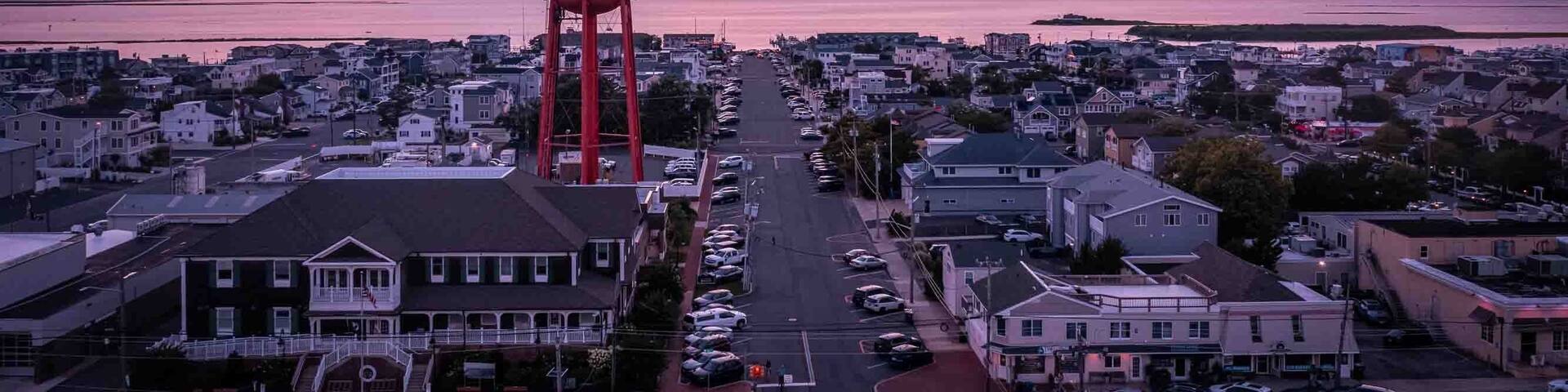 A coastal town at sunset with a prominent red water tower, residential buildings, and a clear view of the ocean.