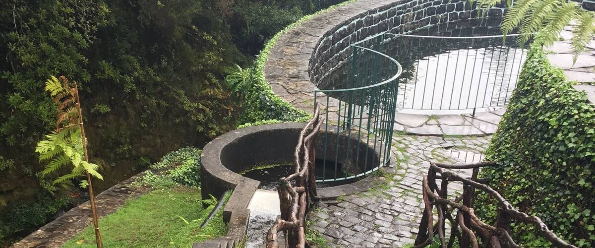 A stone pathway leading to a circular water feature surrounded by greenery and a fence.