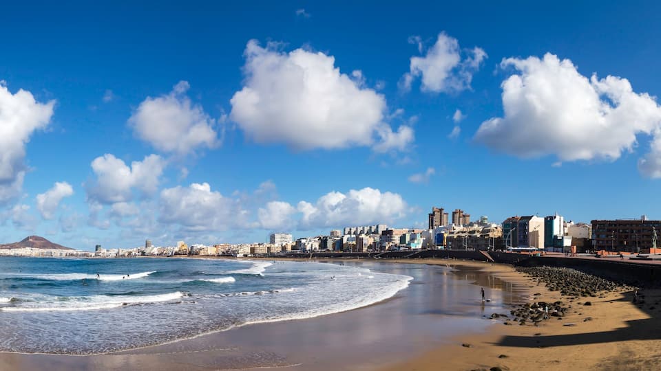 A coastal cityscape with a beach, rocky shoreline, and a clear blue sky with clouds.