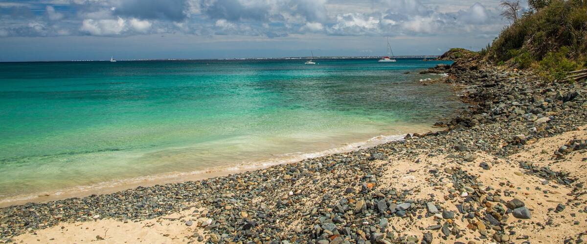 Une plage de galets bordée d’une eau turquoise, avec un voilier visible au loin.