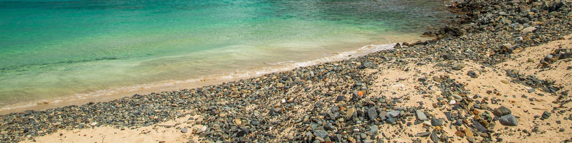 A pebbly beach with turquoise waters and a sailboat in the distance.