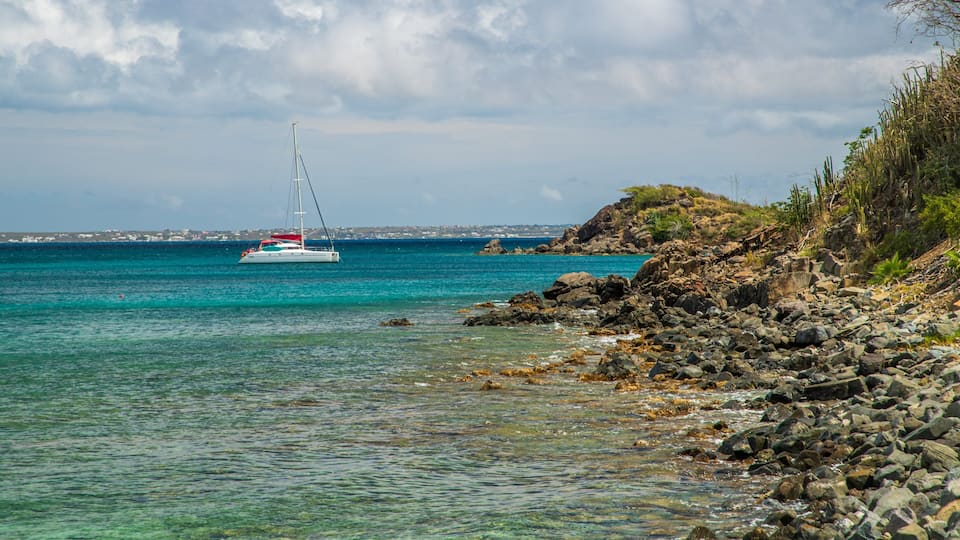 A rocky shoreline with clear turquoise water, a sailboat in the distance, and a coastal town in the background.