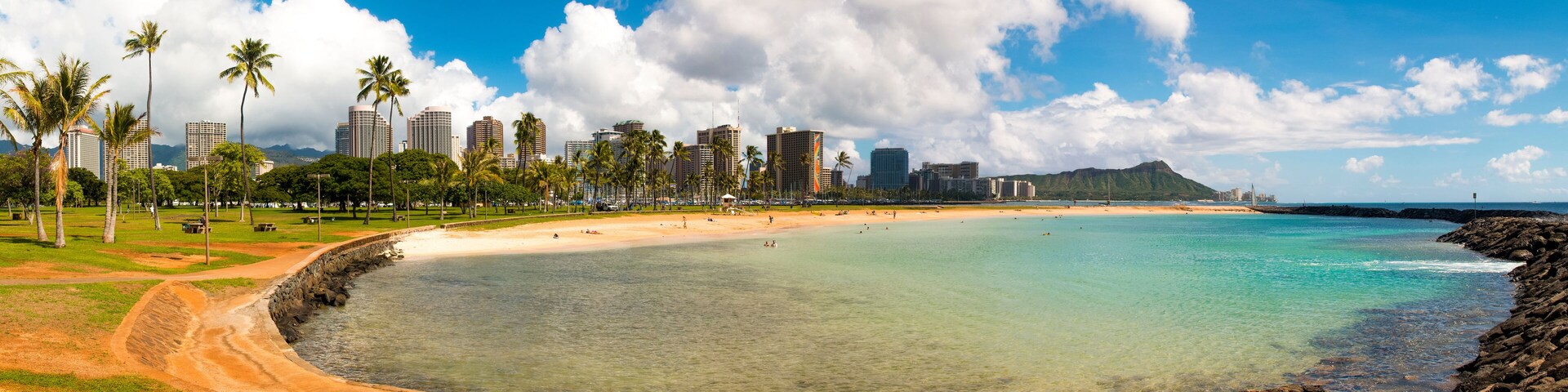 Una playa con agua turquesa cristalina, una orilla arenosa, palmeras y un paisaje urbano con edificios altos al fondo.
