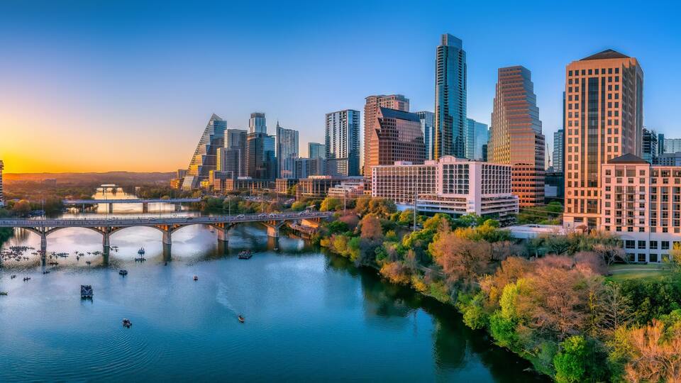 A cityscape with modern skyscrapers, a river, and a bridge during sunset.