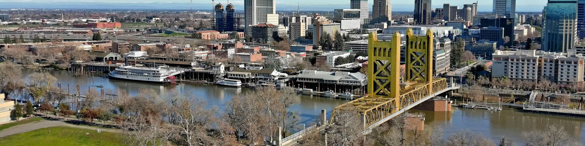 A cityscape with a prominent yellow bridge over a river, surrounded by modern buildings and a clear sky.