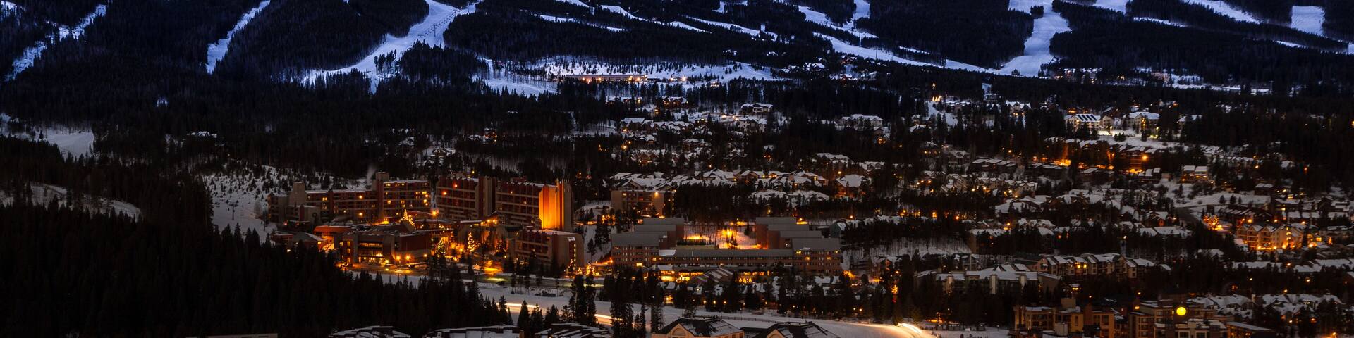 A snow-covered mountain range with a town at its base, illuminated by artificial lights.