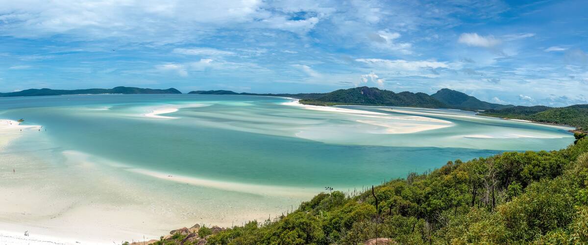 A coastal landscape with clear turquoise waters, white sandy beaches, and lush green hills in the background.
