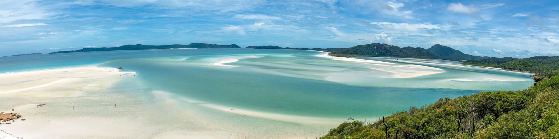 A coastal landscape with clear turquoise waters, white sandy beaches, and lush green hills in the background.