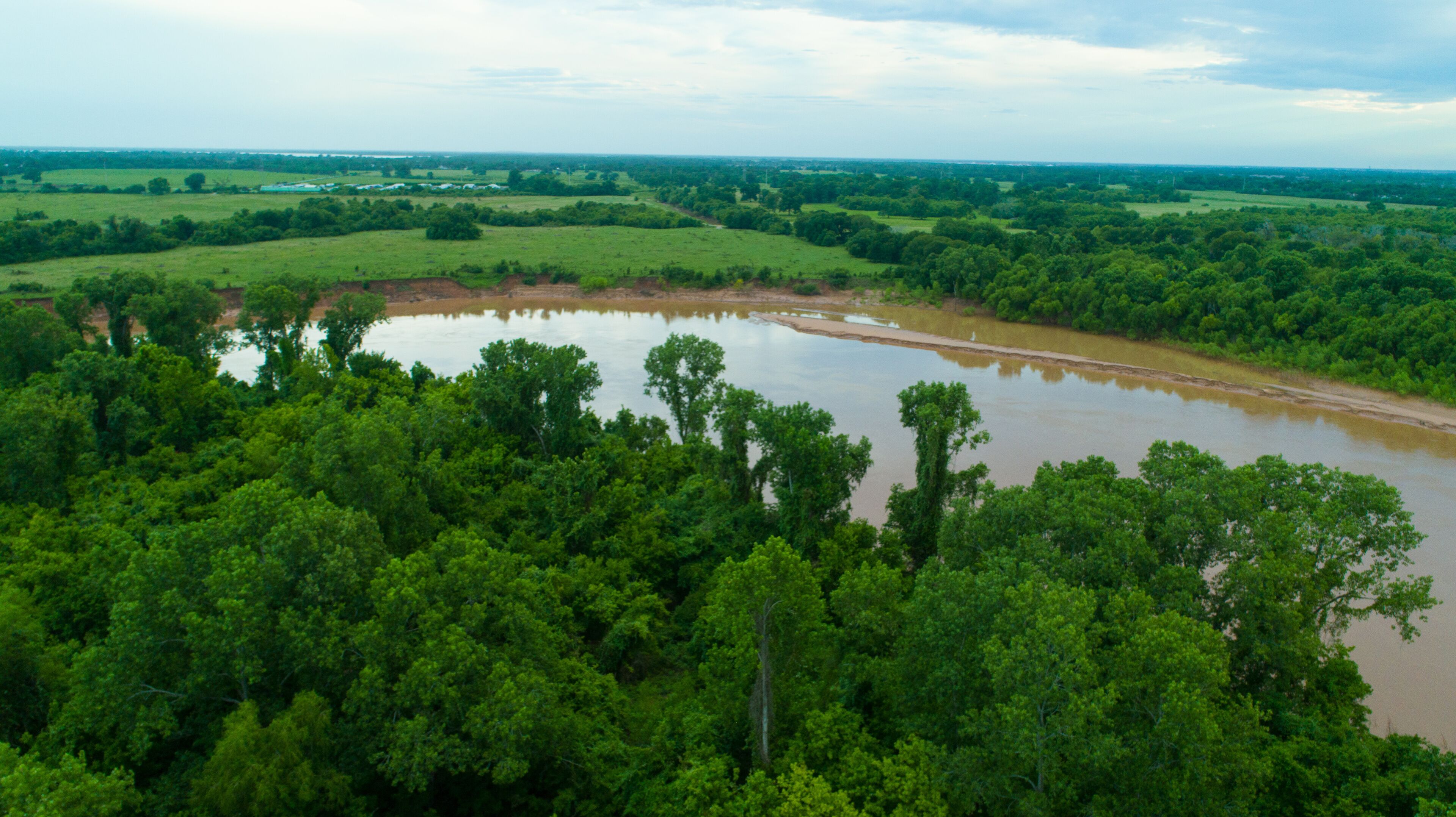 A river winding through a dense forest with a distant view of fields and a cloudy sky.