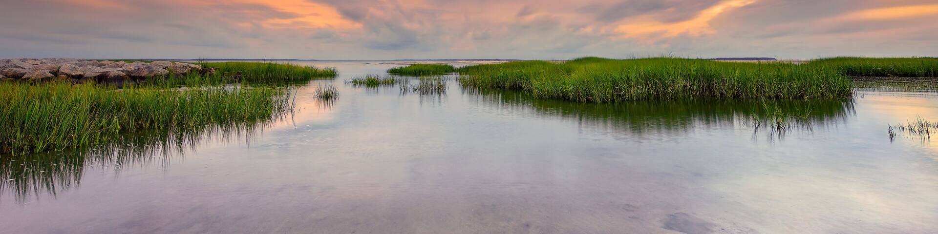 A serene landscape with a calm water body surrounded by green vegetation and a dramatic sky.
