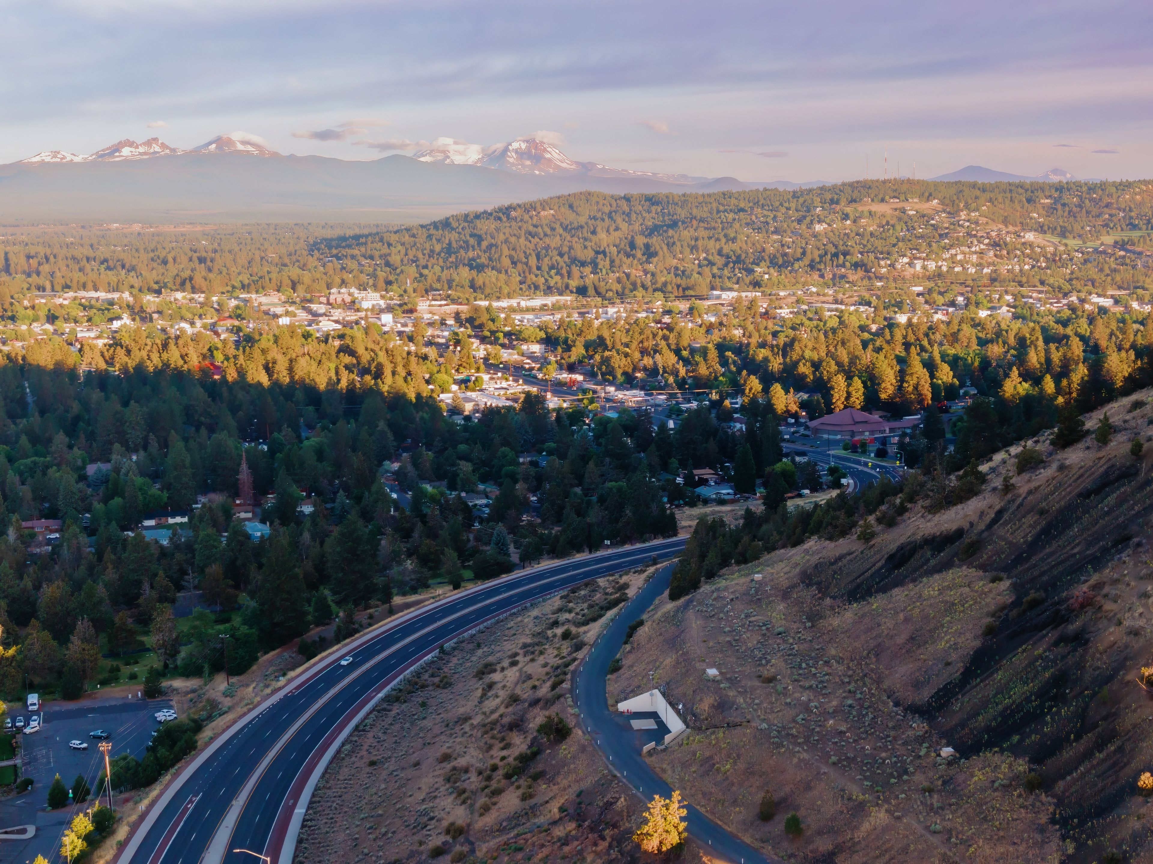 High-angle view of a Bend nestled in a valley, with snow-capped mountains in the background. Highway curves through the landscape. Orchard District, Bend, Oregon, United States