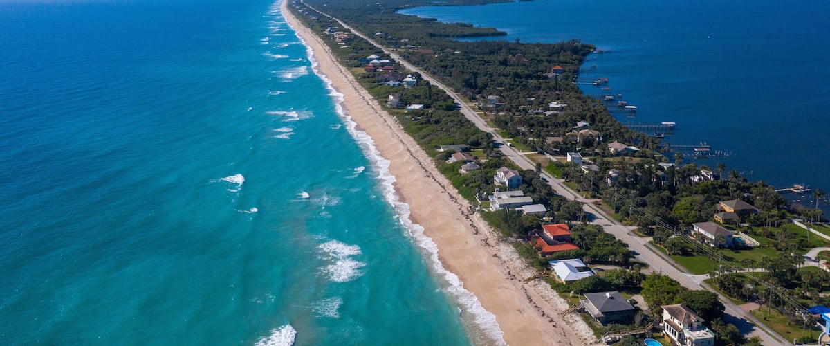 An aerial view of a coastal town with a long beachfront, dense residential area, and clear blue waters.