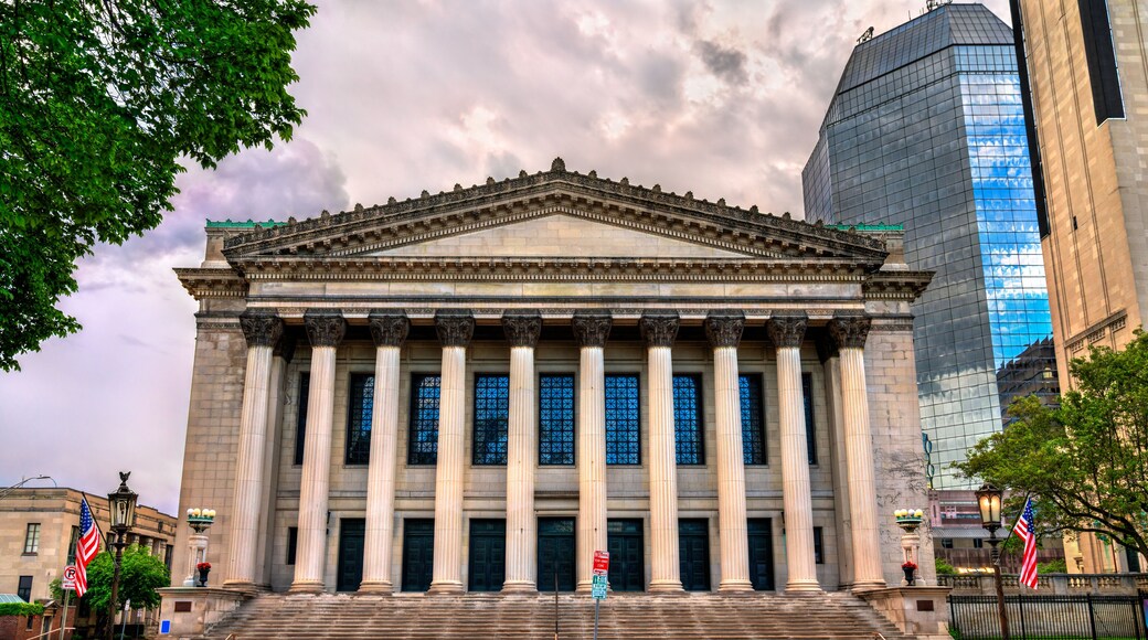 Symphony Hall in Springfield stands with a stately colonnaded facade in Greek Revival style, flanked by American flags and surrounded by a mix of historical and modern architecture. The dramatic cloud