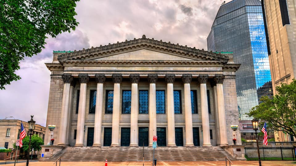 Symphony Hall in Springfield stands with a stately colonnaded facade in Greek Revival style, flanked by American flags and surrounded by a mix of historical and modern architecture. The dramatic cloud