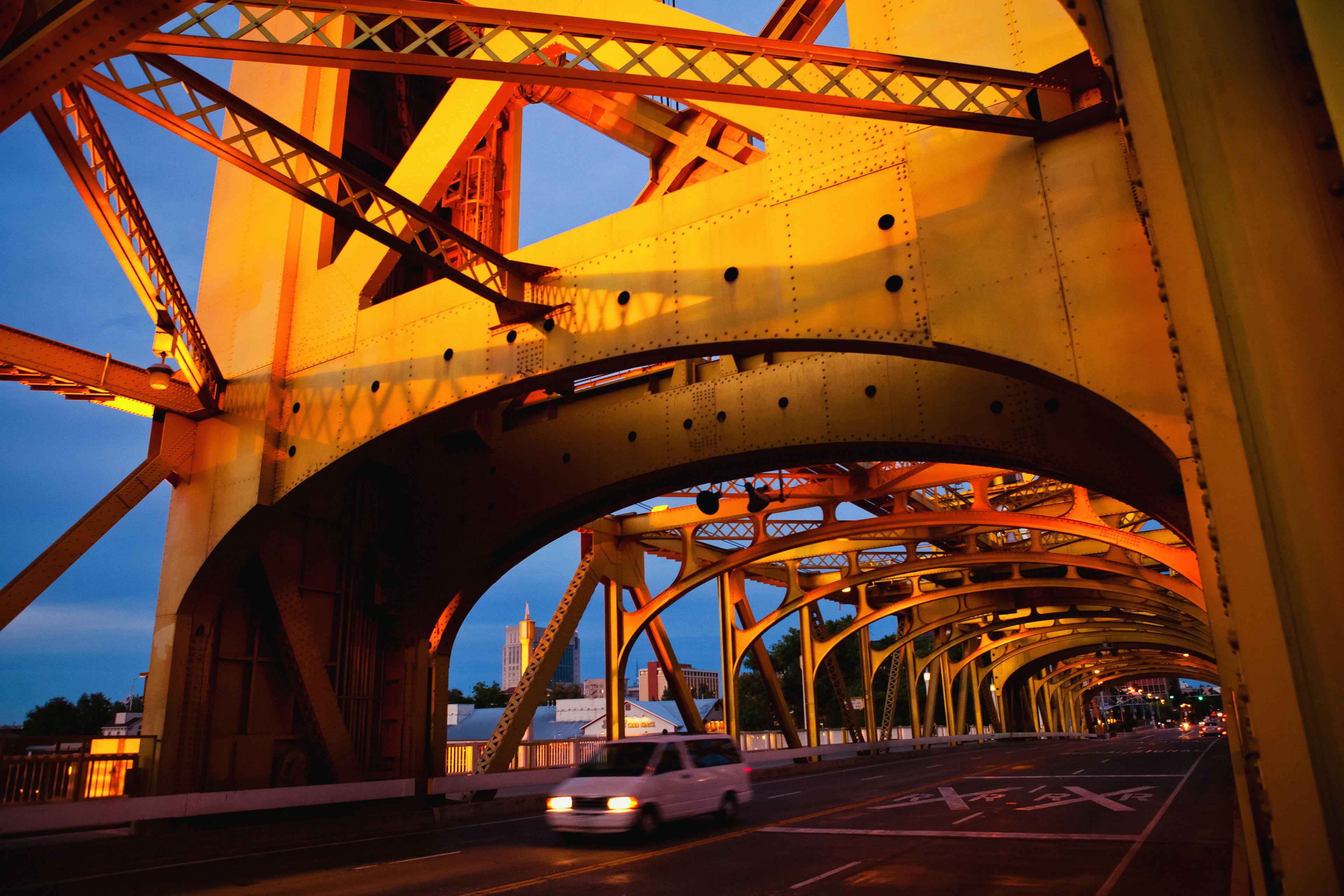 A bridge with yellow trusses and a white car driving on it.