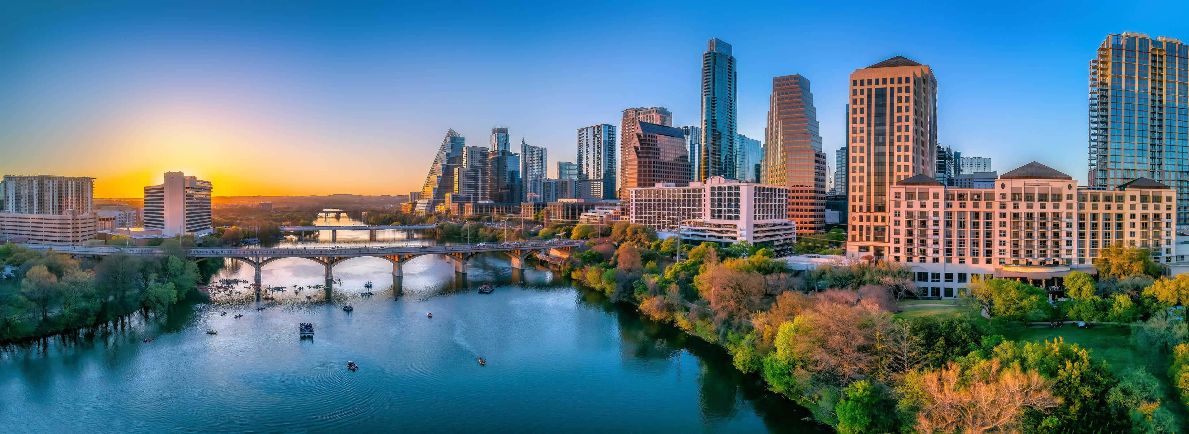 A city skyline with modern skyscrapers, a river with boats, and a bridge.
