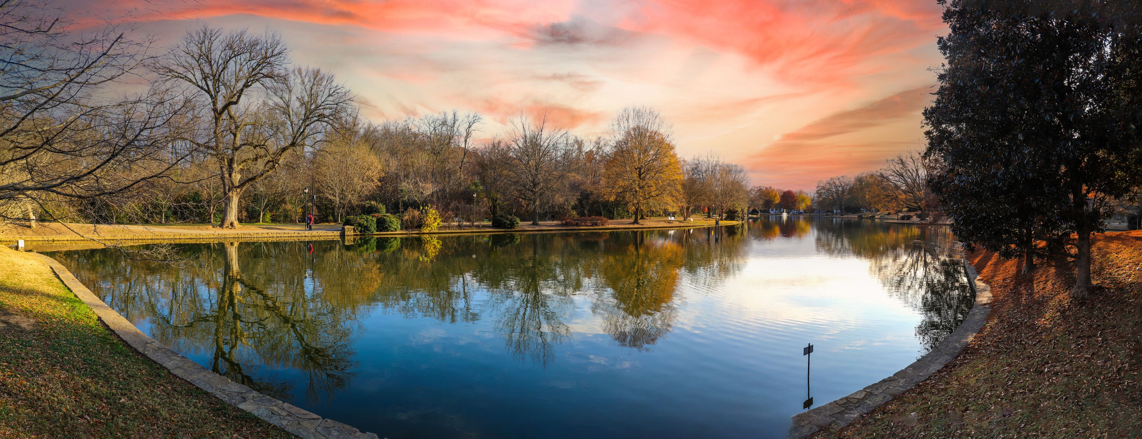 A calm lake with trees reflecting the sky, surrounded by a park with a walking path.