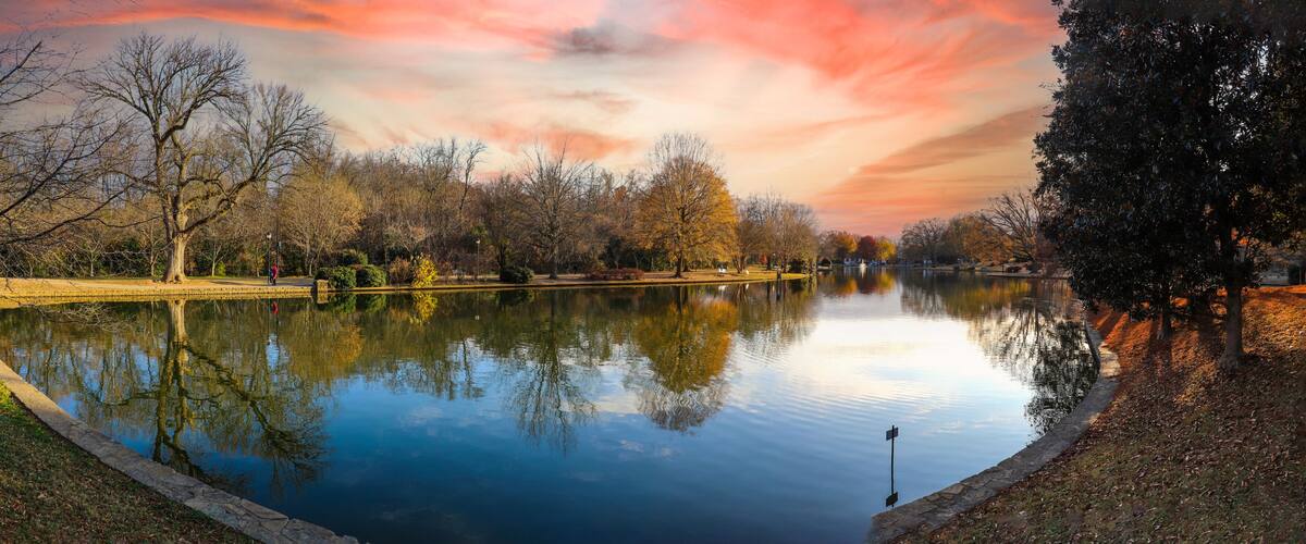 A calm lake with trees reflecting the sky, surrounded by a park with a walking path.