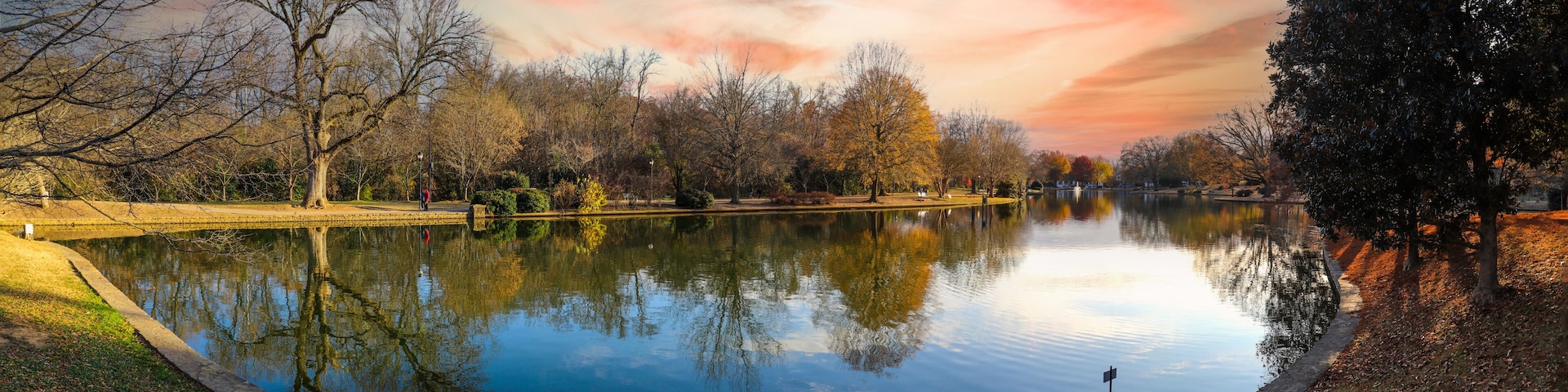 A calm lake with trees reflecting the sky, surrounded by a park with a walking path.