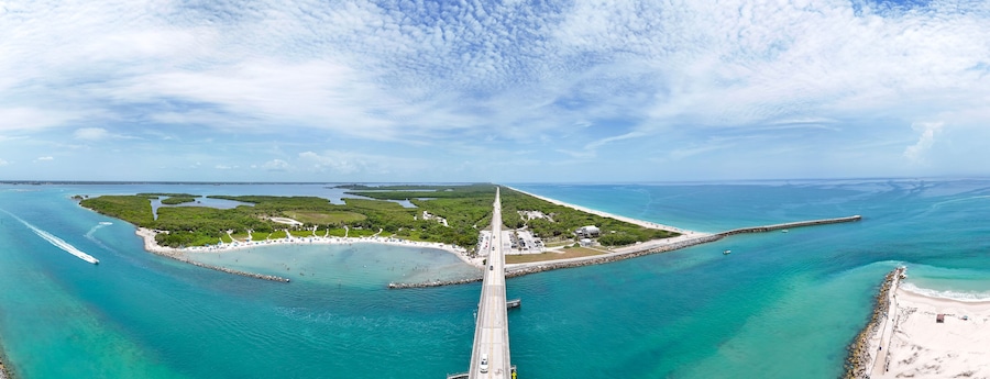 Vista aérea de uma área costeira com uma ponte ligando duas massas de terra, águas turquesa cristalinas e uma praia arenosa.