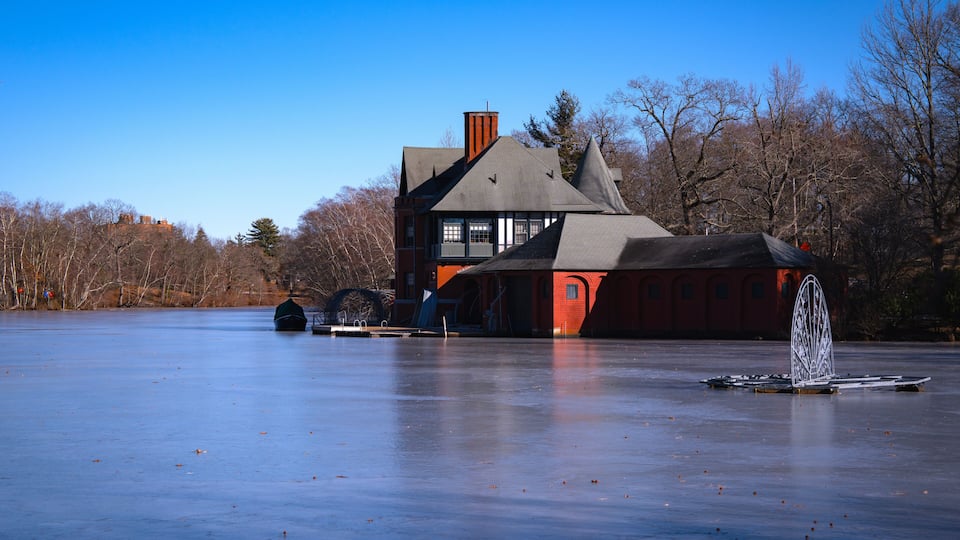 Une grande maison avec un toit en pente et une cheminée, située au bord d’un lac gelé, avec des arbres sans feuilles en arrière-plan.