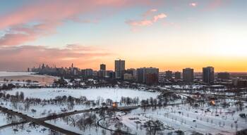 Beautiful Chicago winter skyline aerial above a snow covered Montrose beach looking towards the downtown skyscraper buildings with pink and orange clouds in a blue sky above.